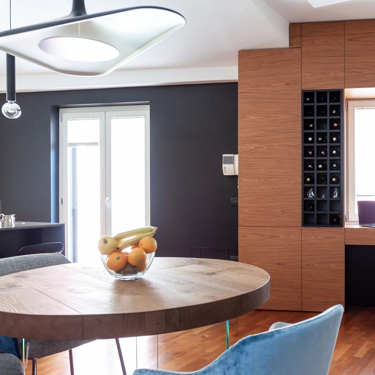 The edge of a white disc pendant glows at the top of a dining corner with a round oak table, a blue chair and a timber cabinet, from Clérachic.