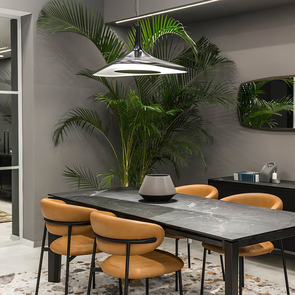 A black disc pendant with a silver-white underside hangs above a dark stone dining table and tan chairs beside a tall palm, from Clérachic.