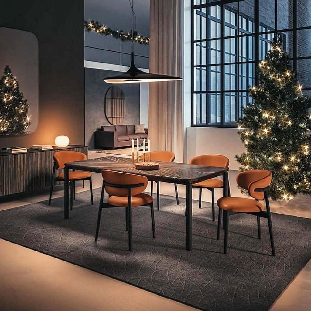A black disc pendant hangs low over a dark dining table with rust-orange chairs, with lit trees and a large factory window behind, from Clérachic.