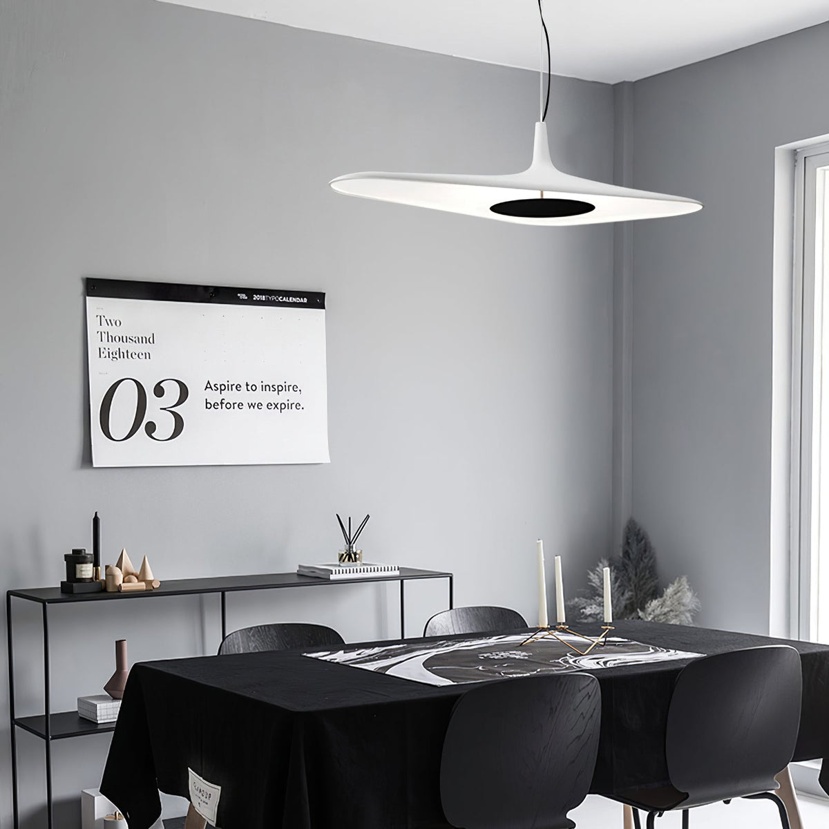A white disc pendant with a dark centre hangs in a grey room above a black table and chairs, with a black console behind, from Clérachic.