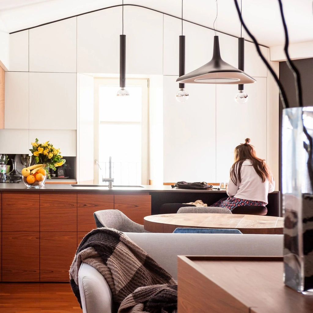 A black and grey disc pendant hangs among slim black tube pendants in a kitchen with warm timber units, in a Clérachic LED pendant setting.