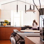 A black and grey disc pendant hangs among slim black tube pendants in a kitchen with warm timber units, in a Clérachic LED pendant setting.