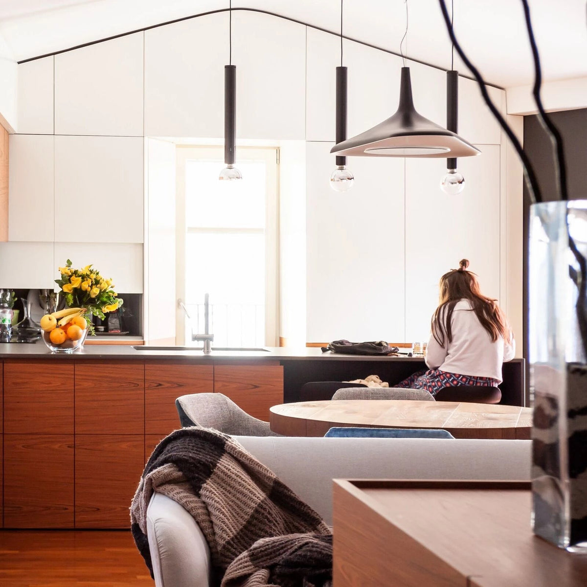 A black and grey disc pendant hangs among slim black tube pendants in a kitchen with warm timber units, in a Clérachic LED pendant setting.