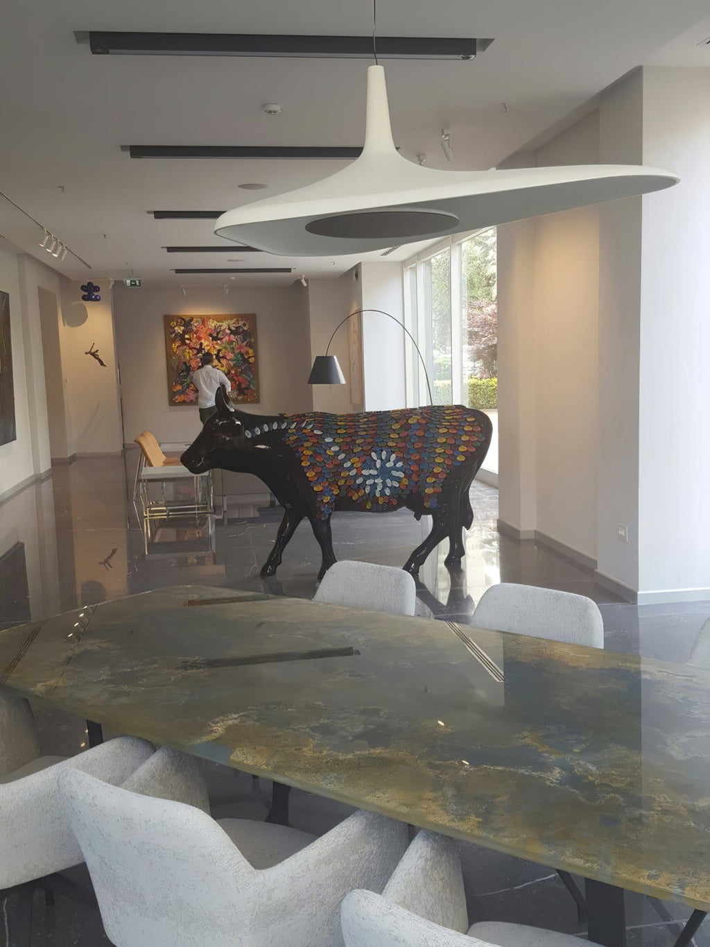 A white disc pendant seen from close below hangs in a long gallery room with a patterned cow sculpture and a stone-topped table, from Clérachic.
