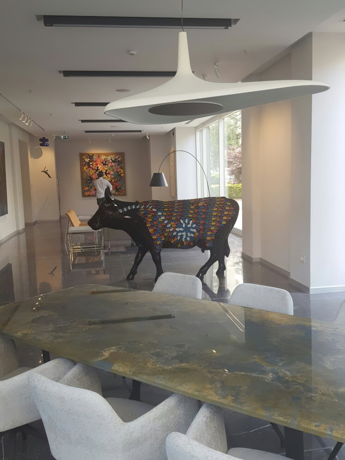 A white disc pendant seen from close below hangs in a long gallery room with a patterned cow sculpture and a stone-topped table, from Clérachic.