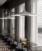 Two white disc pendants hang over a long row of dark chairs and glass tables beside tall windows, in a Clérachic LED pendant setting.