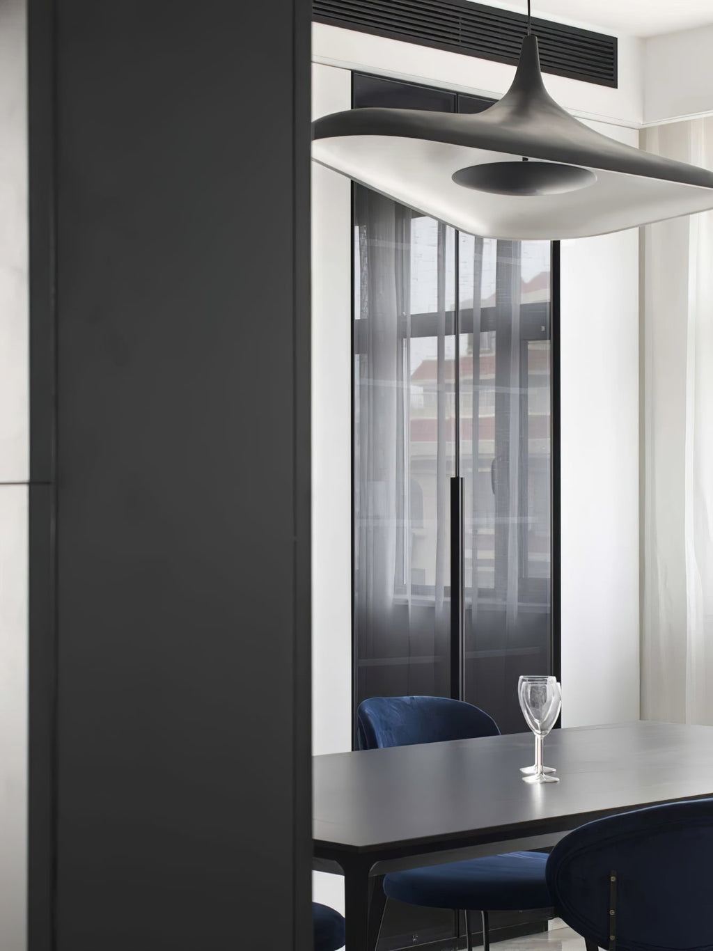 A white disc pendant fills the top of the frame above a black dining table with navy chairs and a wine glass, from Clérachic.