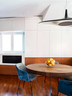 A black pendant with a pale underside enters the frame beside a white wall of cabinets, above a round wooden table and blue chair, from Clérachic.