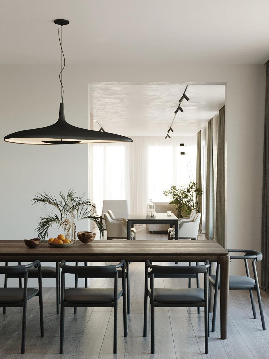 A black disc pendant hangs over a long dark wood dining table with black chairs, with a plant and a sunlit room behind, from Clérachic.