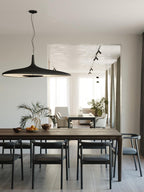 A black disc pendant hangs over a long dark wood dining table with black chairs, with a plant and a sunlit room behind, from Clérachic.