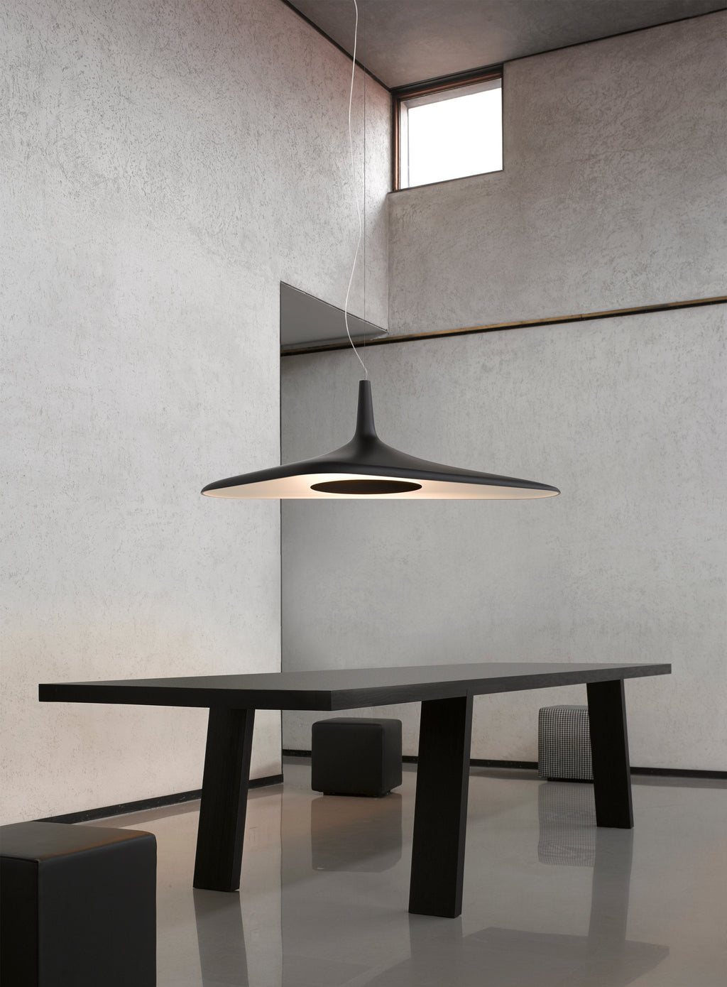 A black disc pendant hangs over a black dining table in a tall room of rough grey plaster with a high window, a Clérachic LED pendant.