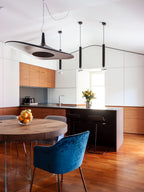 A black disc pendant with a dark centre hangs in a kitchen with timber cabinets, next to slim black tube pendants, from Clérachic.