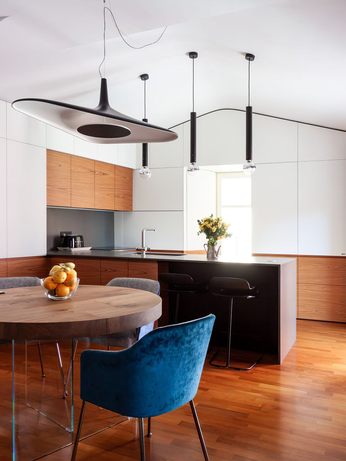 A black disc pendant with a dark centre hangs in a kitchen with timber cabinets, next to slim black tube pendants, from Clérachic.