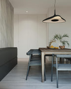 A black disc pendant with a lit white inner surface hangs above a timber dining table in a pale room with a palm in a bowl, from Clérachic.