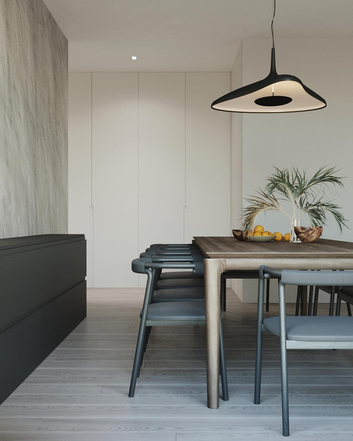 A black disc pendant with a lit white inner surface hangs above a timber dining table in a pale room with a palm in a bowl, from Clérachic.
