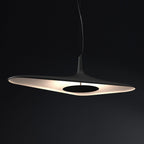 A black disc pendant glows with warm light on its underside against a dark black backdrop, shown as this Clérachic LED pendant.