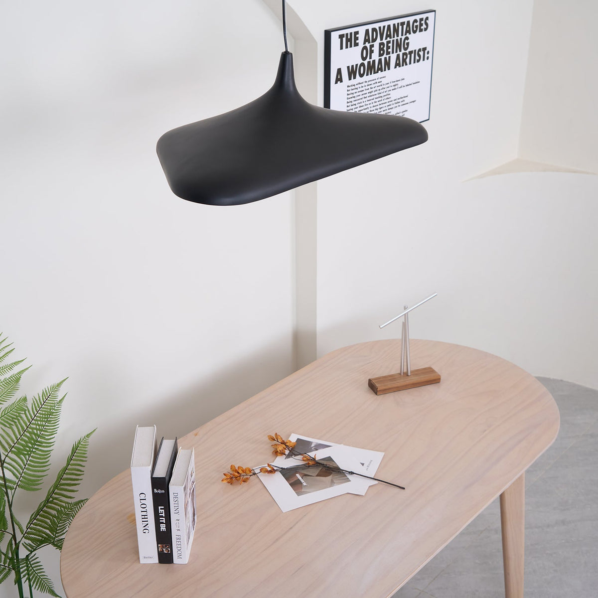 A black disc pendant hangs close above a pale oak oval table with books and dried stems, beside a printed poster, from Clérachic.
