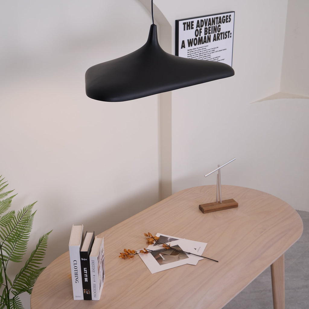 A black disc pendant seen from below left hangs above a pale oak table with books and dried flowers, a Clérachic LED pendant.