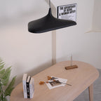 A black disc pendant seen from below left hangs above a pale oak table with books and dried flowers, a Clérachic LED pendant.