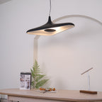 A black disc pendant lit warm on its underside hangs in front of a white arch above a pale table with a fern, from Clérachic.