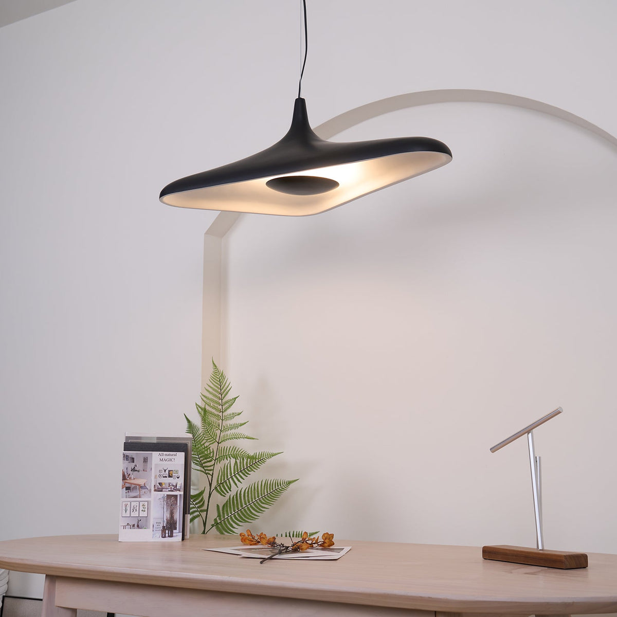 A black disc pendant lit warm on its underside hangs in front of a white arch above a pale table with a fern, from Clérachic.