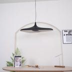 A black disc pendant, switched off, hangs in front of a white arch above a pale oak table with a book and a fern, from Clérachic.