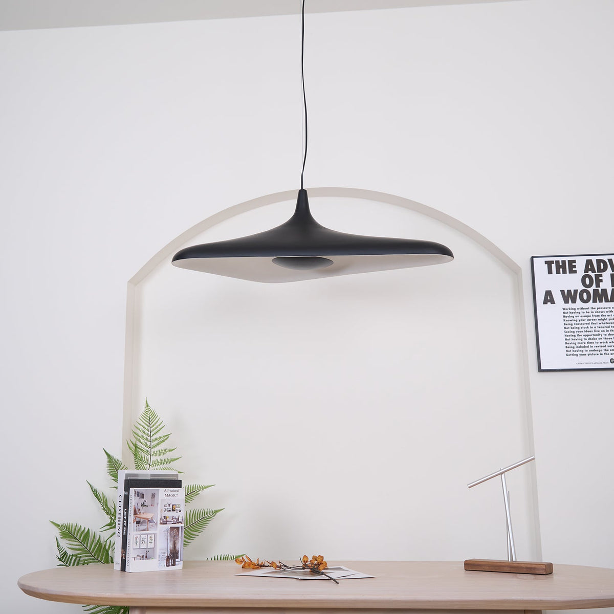 A black disc pendant, switched off, hangs in front of a white arch above a pale oak table with a book and a fern, from Clérachic.