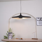 The same black disc pendant switched on, its pale inner surface glowing warm in front of a white arch, from Clérachic.