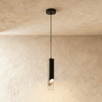 Clérachic single black cylinder pendant with an angled cut and clear glass tip, glowing warmly on a thin black cord.