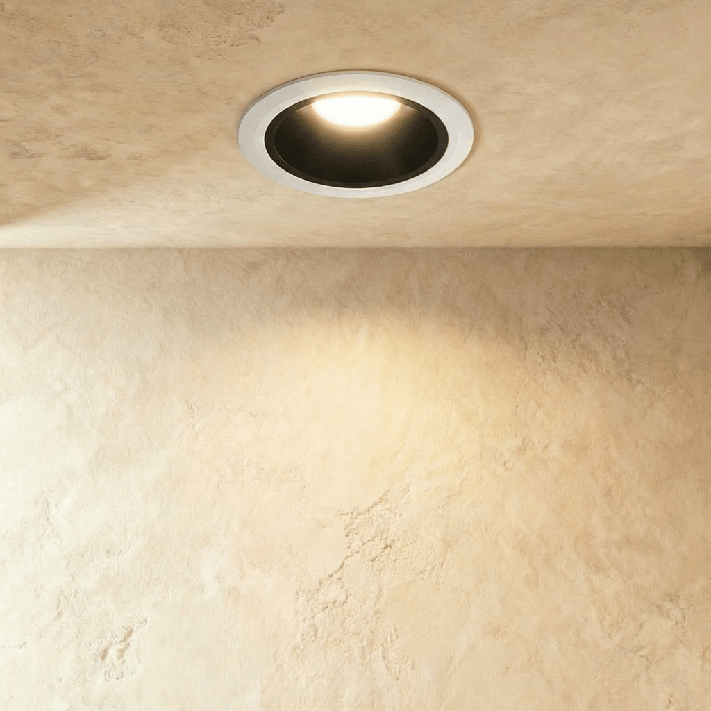 Clérachic recessed downlight with a white trim and black inner cone, lit warm white against a sand-coloured plaster wall.