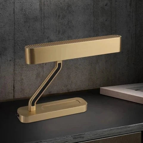 Clérachic brushed brass table lamp with a slim grilled head and angled arm, set on a dark shelf against a grey textured wall.