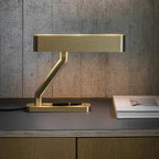 Clérachic brass table lamp glowing warmly along its slotted head, on a desk with a closed pen and a book against concrete.