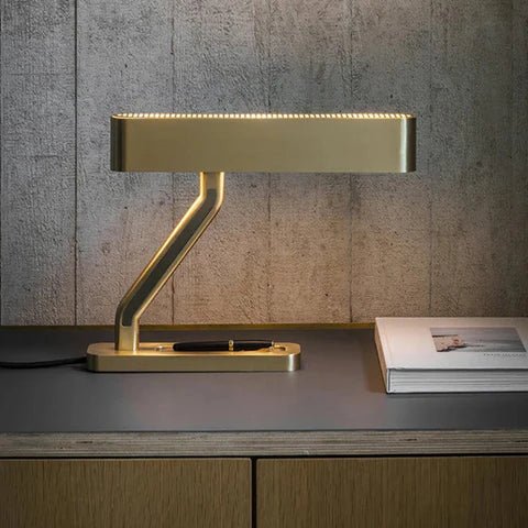Clérachic brass table lamp glowing warmly along its slotted head, on a desk with a closed pen and a book against concrete.