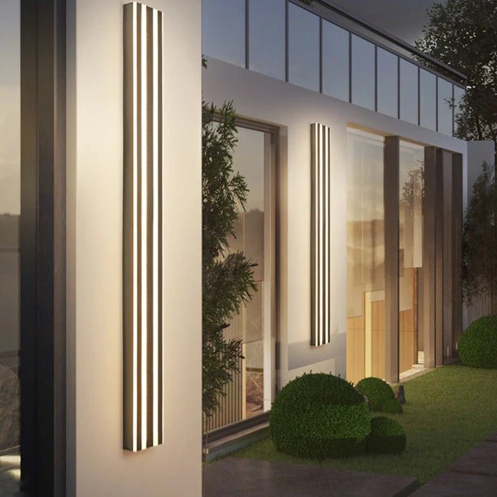 Two Clérachic long dark fluted LED bar wall lights glowing at dusk on a white modern facade beside clipped garden hedges.