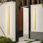 Two Clérachic gold fluted LED bar wall lights glowing on white courtyard walls either side of a black-framed gateway.