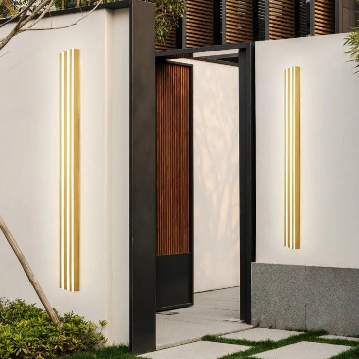 Two Clérachic gold fluted LED bar wall lights glowing on white courtyard walls either side of a black-framed gateway.