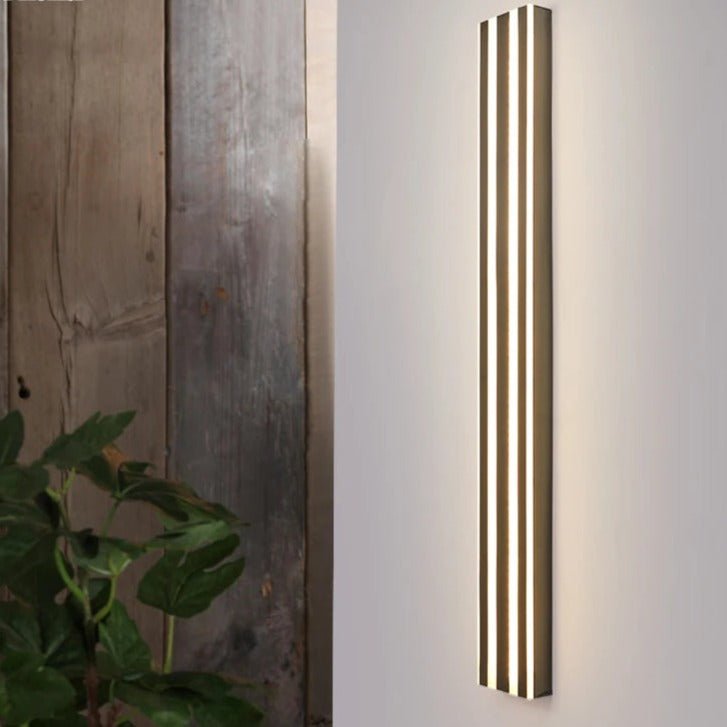 Clérachic tall dark fluted LED bar wall light lit on a pale wall beside weathered timber boards and a leafy plant.