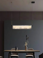 Clérachic Linear Alabaster LED Pendant Light against a deep green-grey wall, the veined stone lit above a dining table set with brass reeds.