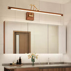 Lit Clérachic linear wall light above a mirrored cabinet in a bathroom, with a wooden door reflected and flowers on the vanity.