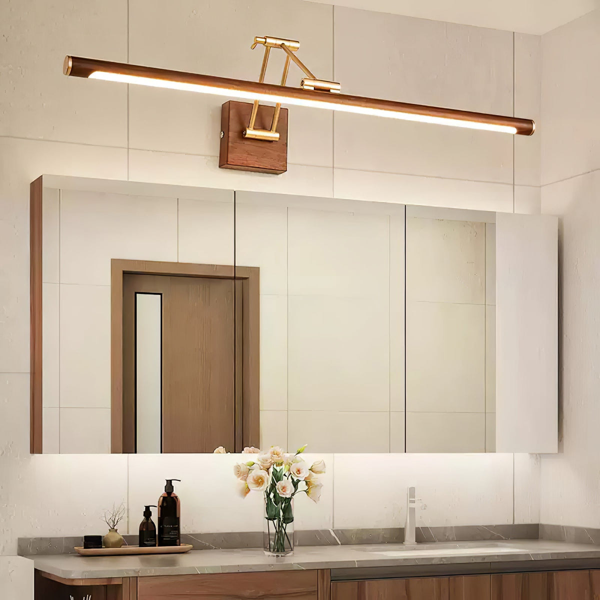 Lit Clérachic linear wall light above a mirrored cabinet in a bathroom, with a wooden door reflected and flowers on the vanity.