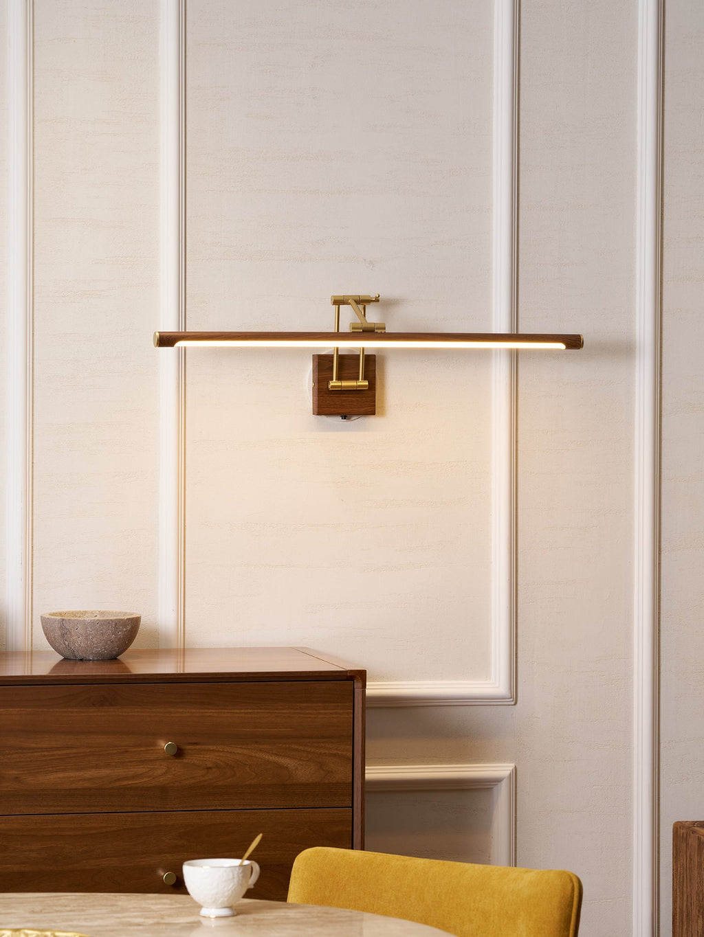 This Clérachic linear wall light lit above a walnut chest of drawers, in a room with moulded panels, a stone bowl and a yellow chair.