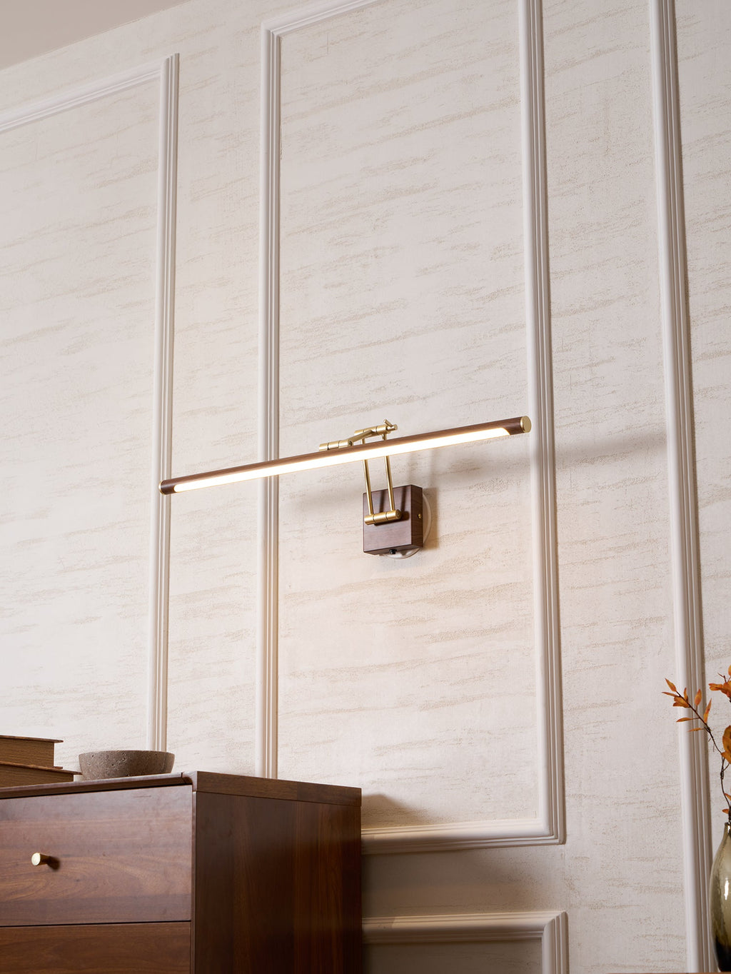 A Clérachic linear wall light lit and tilted on a panelled wall above a walnut chest, with orange leaves at the edge of the frame.