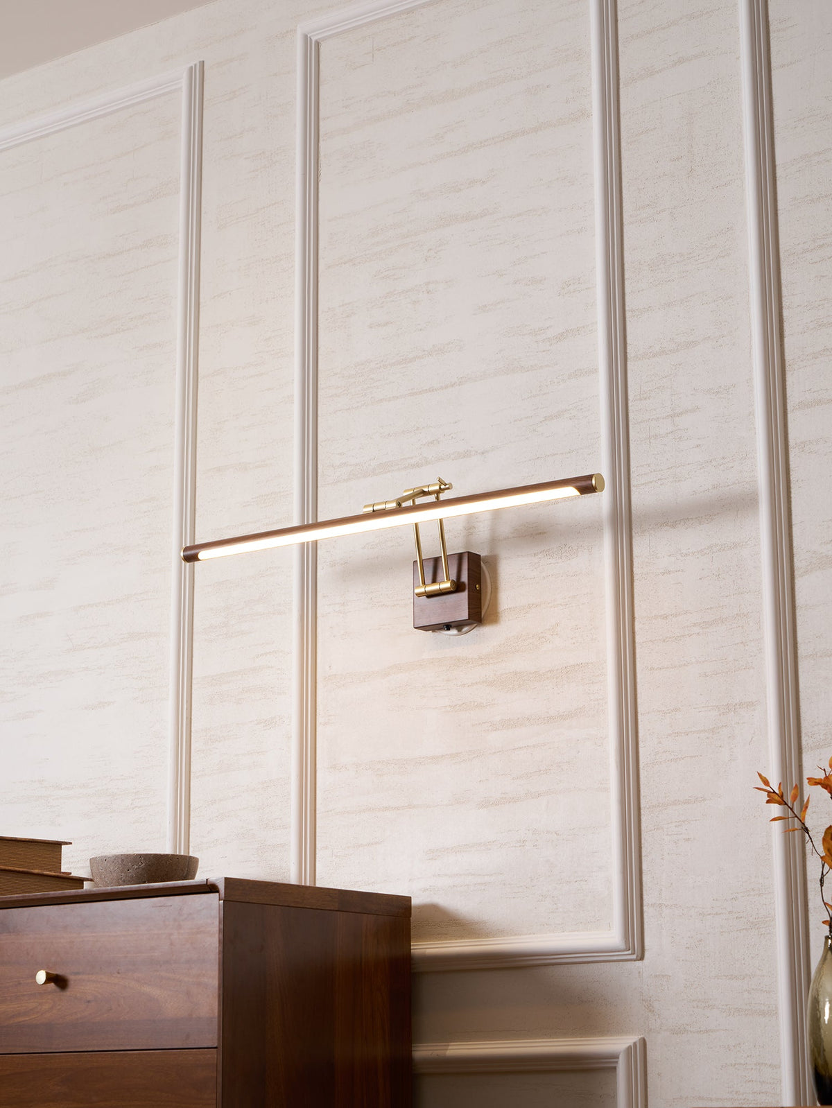 A Clérachic linear wall light lit and tilted on a panelled wall above a walnut chest, with orange leaves at the edge of the frame.