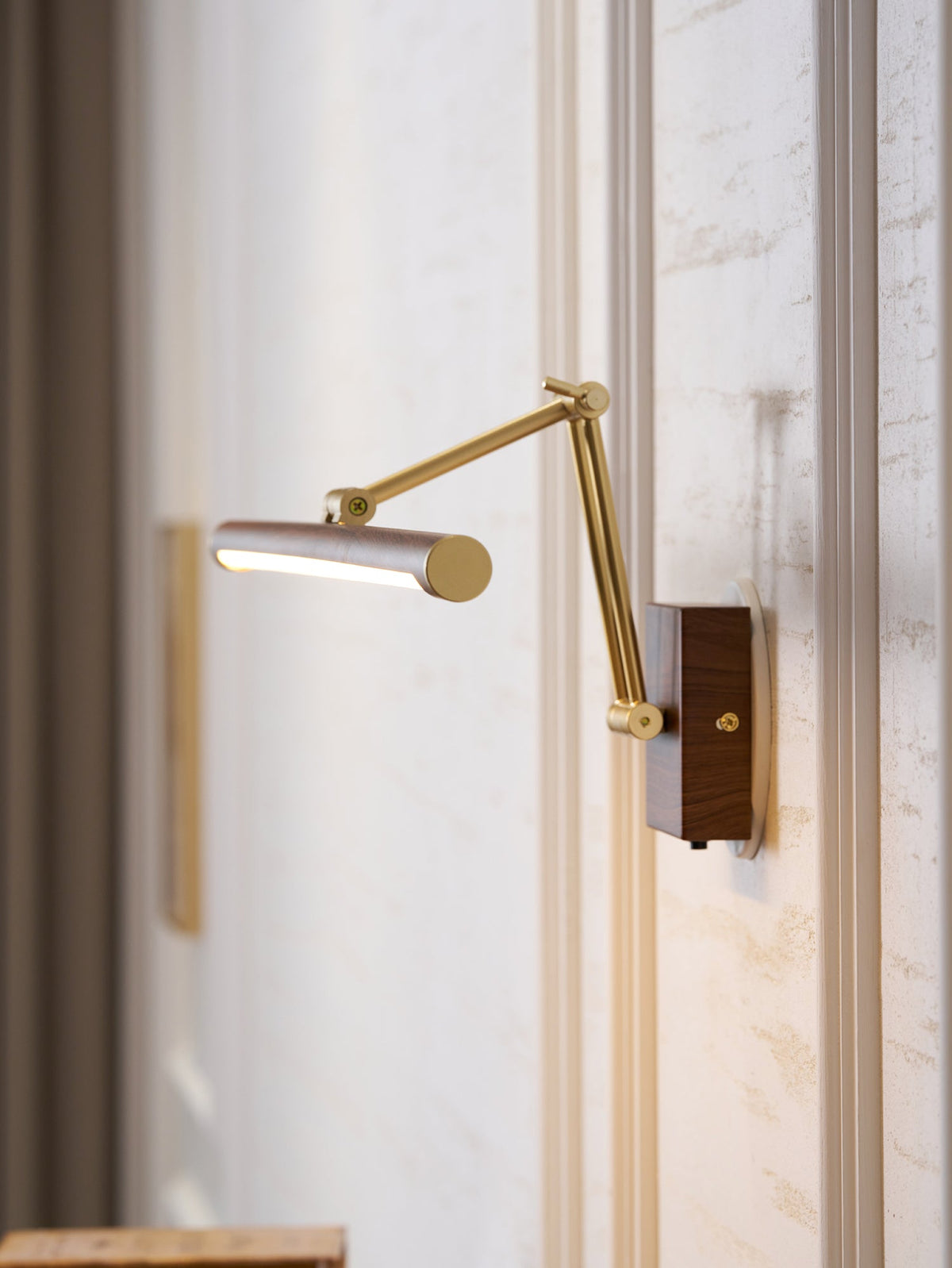 Side-on view of this Clérachic wall light, the lit tube angled forward on a folding brass arm beside a dark curtain.