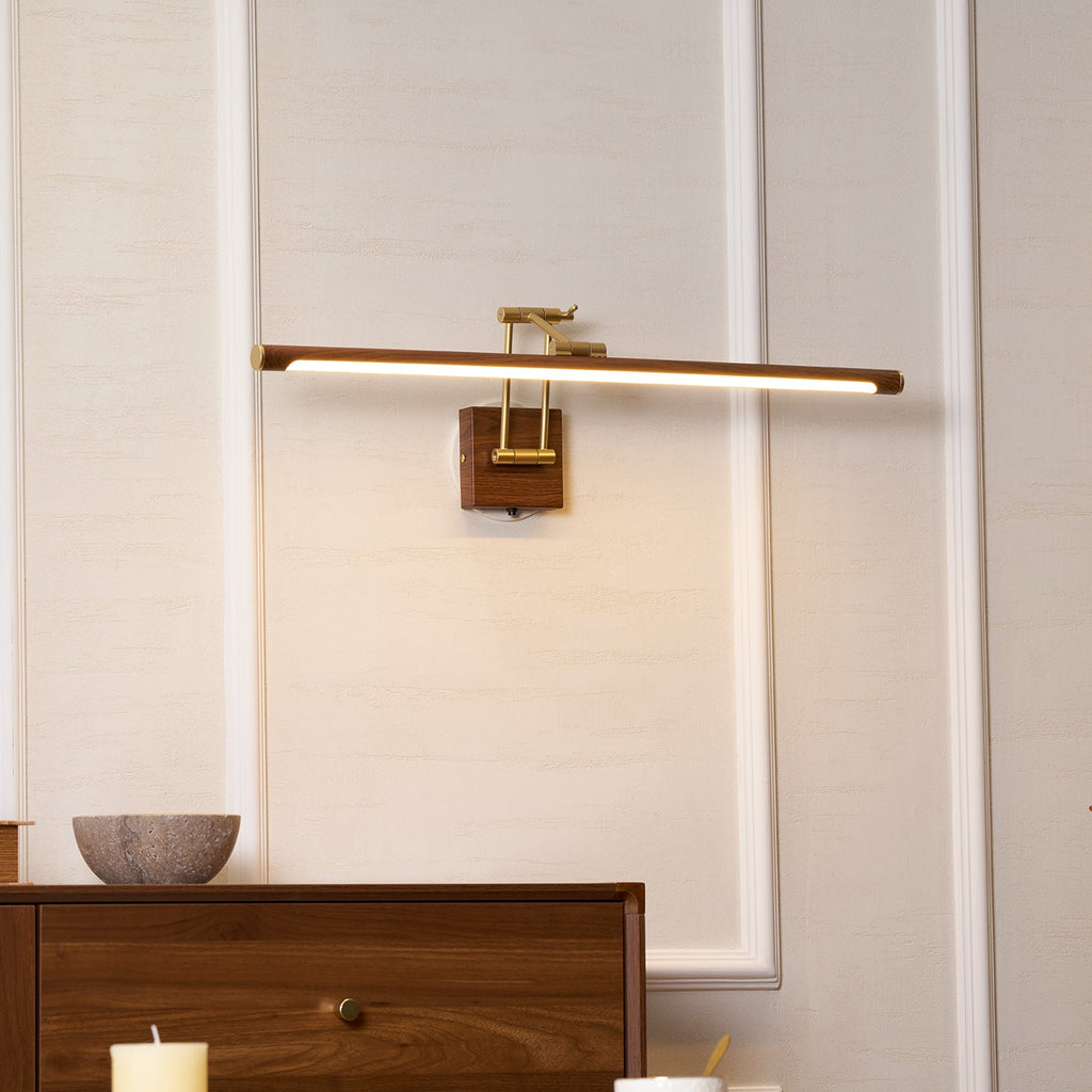 Lit above a walnut chest of drawers and a stone bowl, this Clérachic linear wall light hangs on a panelled wall with a candle below.