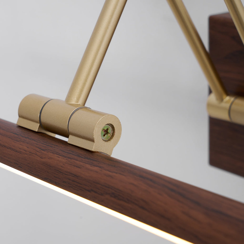 Close view of a brass hinge fixed with a screw to the walnut tube of this Clérachic wall light, the lit strip glowing below.
