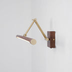 This Clérachic wall light with its brass arm extended and the walnut tube turned end-on, lit, against a pale grey corner.