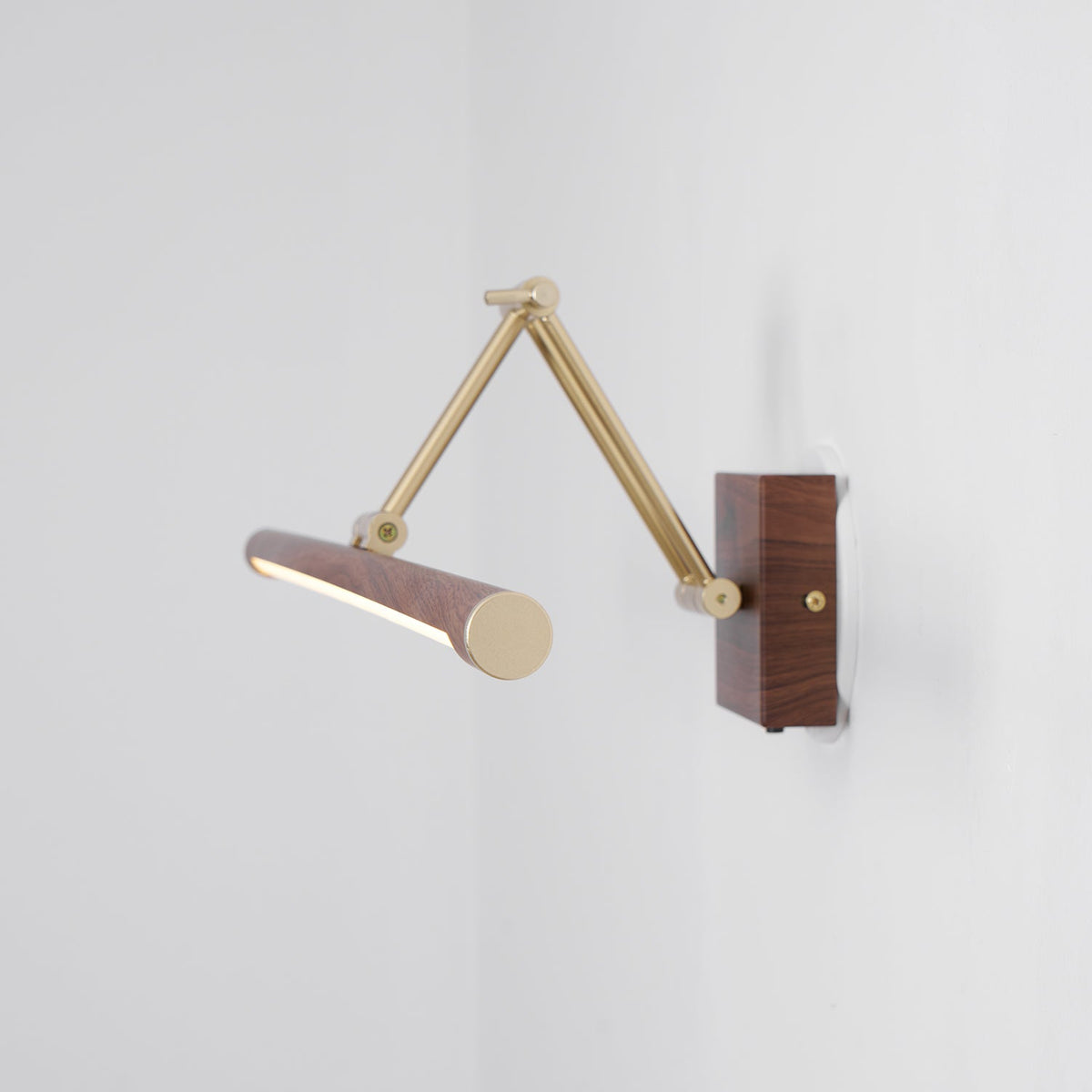 This Clérachic wall light with its brass arm extended and the walnut tube turned end-on, lit, against a pale grey corner.