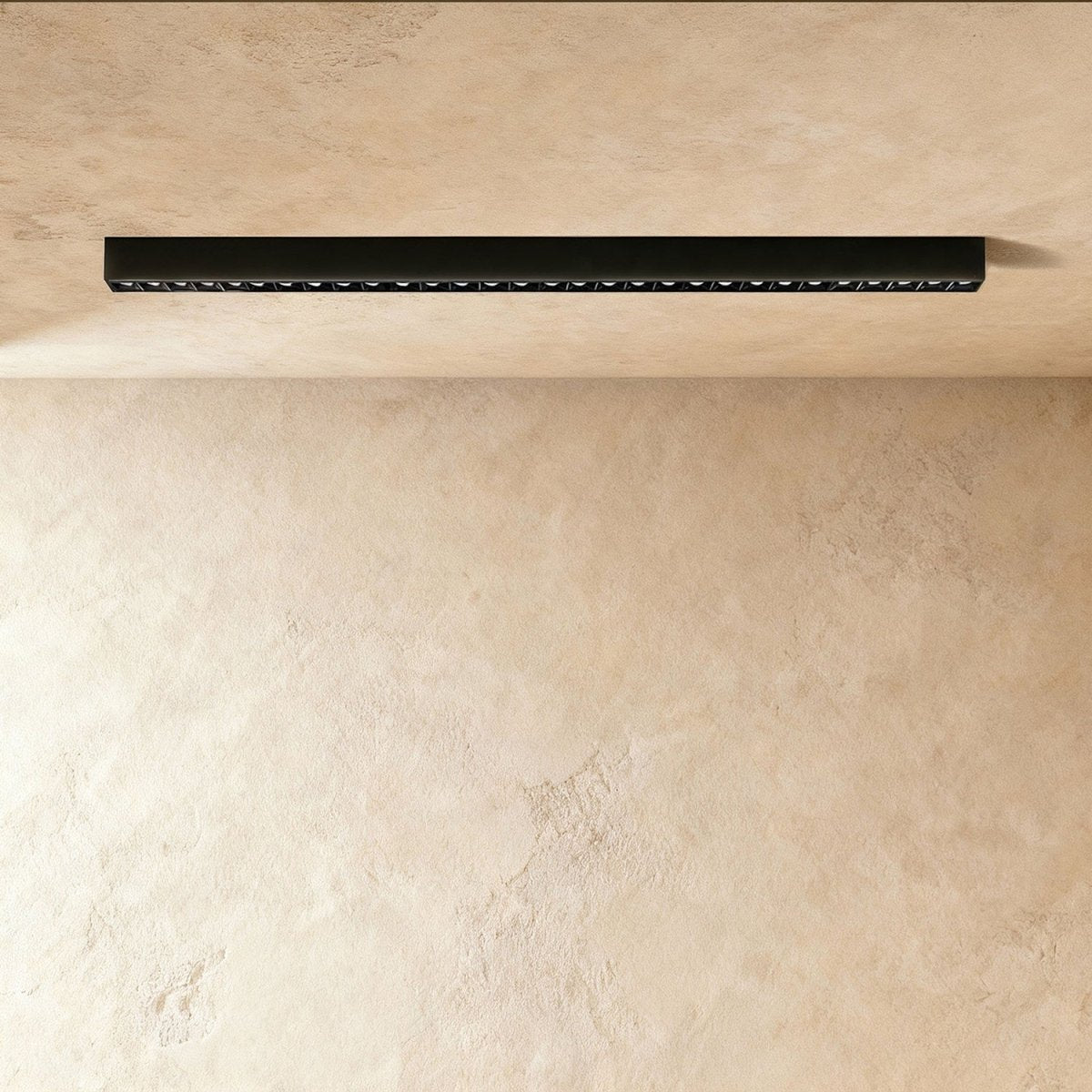 A black linear LED bar with a row of floodlight lenses sits flush in a ceiling corner above a warm plaster wall, a Clérachic fixture.