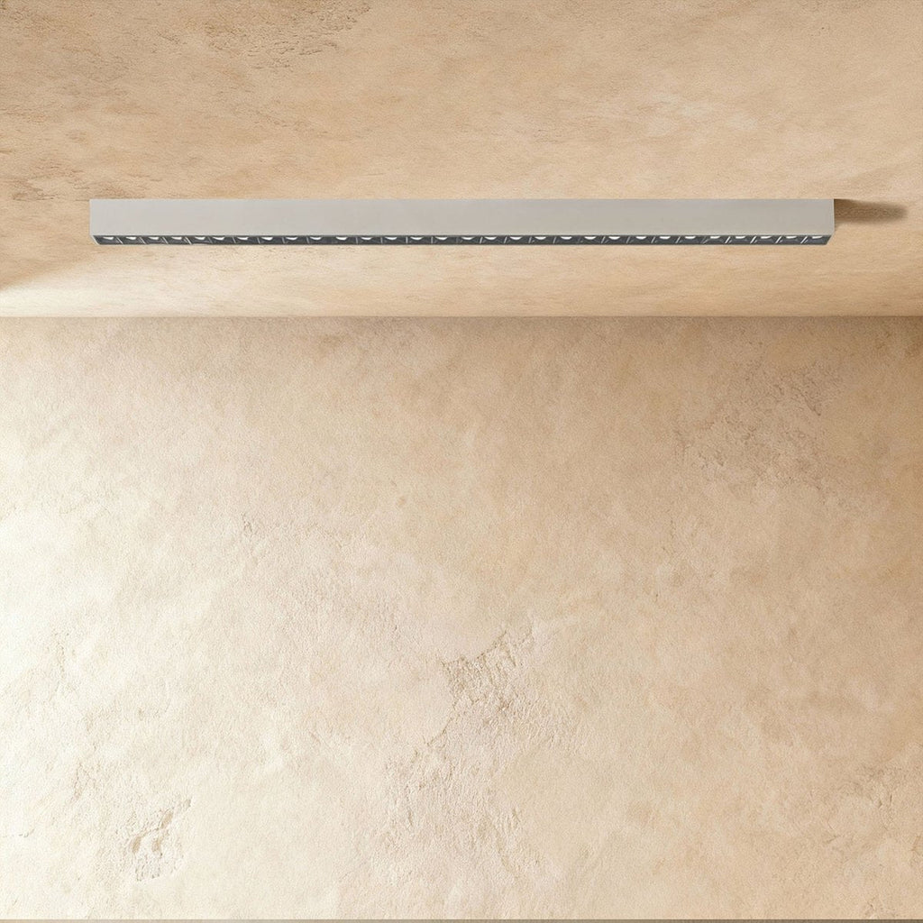 A pale silver-white linear LED bar with a row of floodlight lenses sits flush in a ceiling corner, this Clérachic fixture unlit.